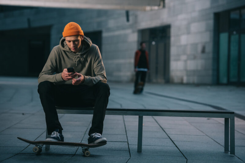 Skateboarder in modern streetwear sitting on bench and using smartphone Historia polskiego streetwearu. Początki streetwearu w Polsce, znaczenie streetwearu dla polskiej mody
